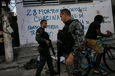 Bereitschaftspolizisten patrouillieren in Rio De Janeiro. Auf der Mauer hinter ihnen steht in portugiesischer Sprache: "28 Tote sind ein Massaker! Unsere Toten haben eine Stimme! Ein Jahr Jacarezinho-Massaker".