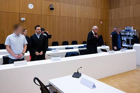 Im Hochsicherheitsgerichtssaal vom Landgericht München II stehen die zwei wegen Mordes angeklagten Männer (l und r) neben ihren Rechtsanwälten.