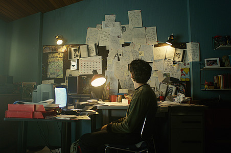 Ein Junge sitzt mit dem Gesicht zu einem dunklen Schreibtisch, über dem viele Zettel und Pläne an der Wand hängen.