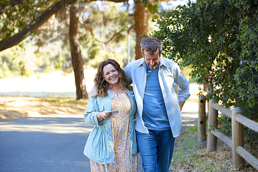 Melissa McCarthy und Chris O'Dowd spazieren lachend und Händchen haltend durch einen Park.